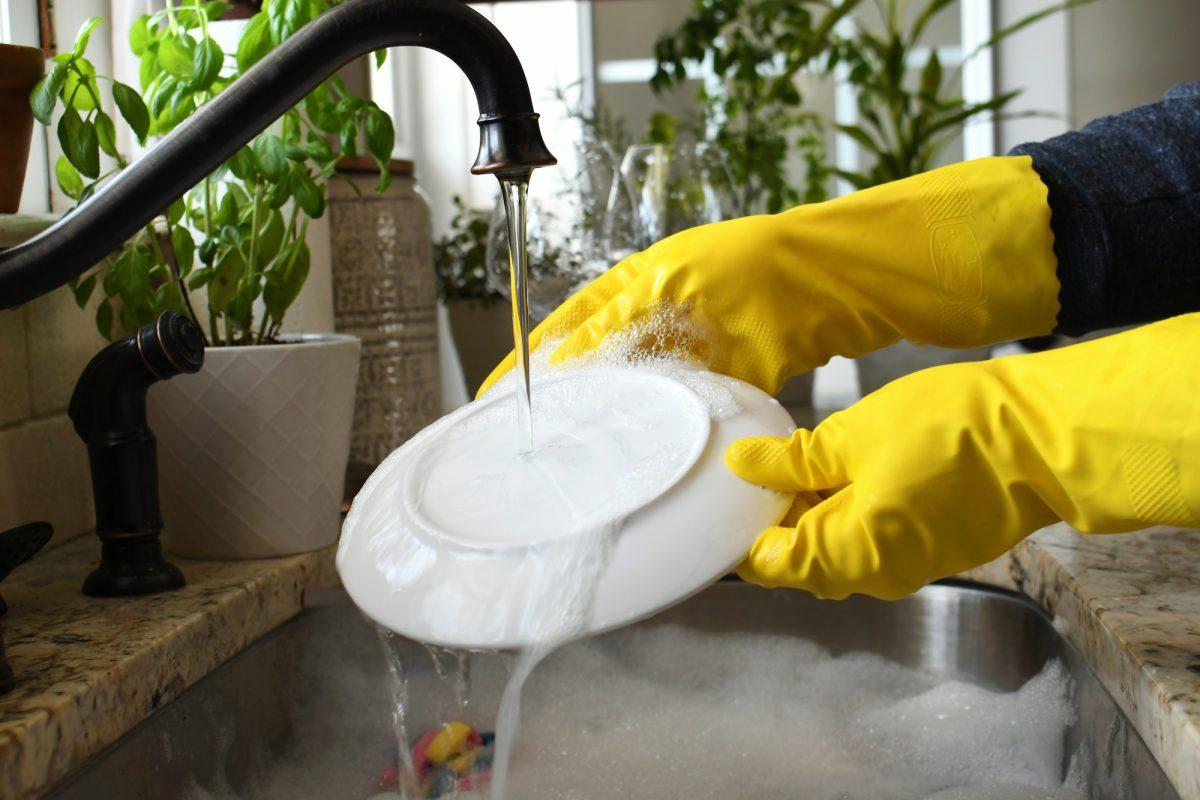 Washing rinsing dishes in kitchen sink by hand wearing yellow rubber gloves. running water