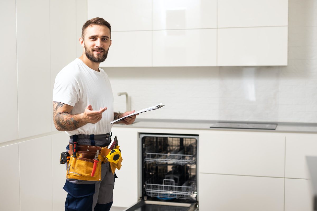 Young modern serviceman in worker suit during the repairing of the dishwasher on the domestic